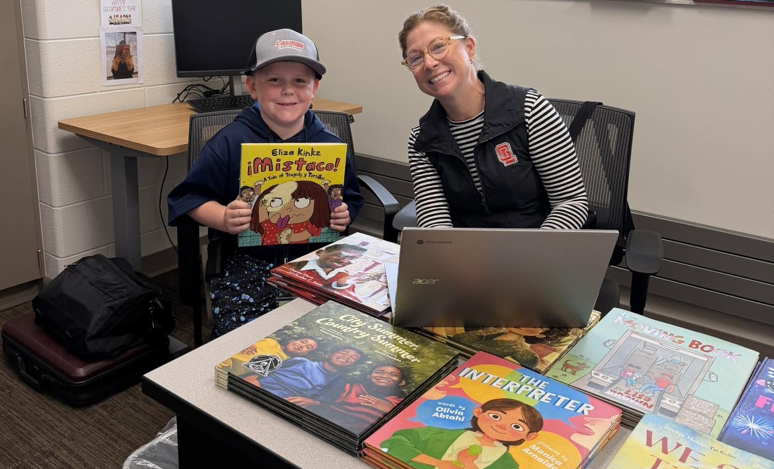 A student holding up a new picture book beside a teacher, with stacks of new books on the table