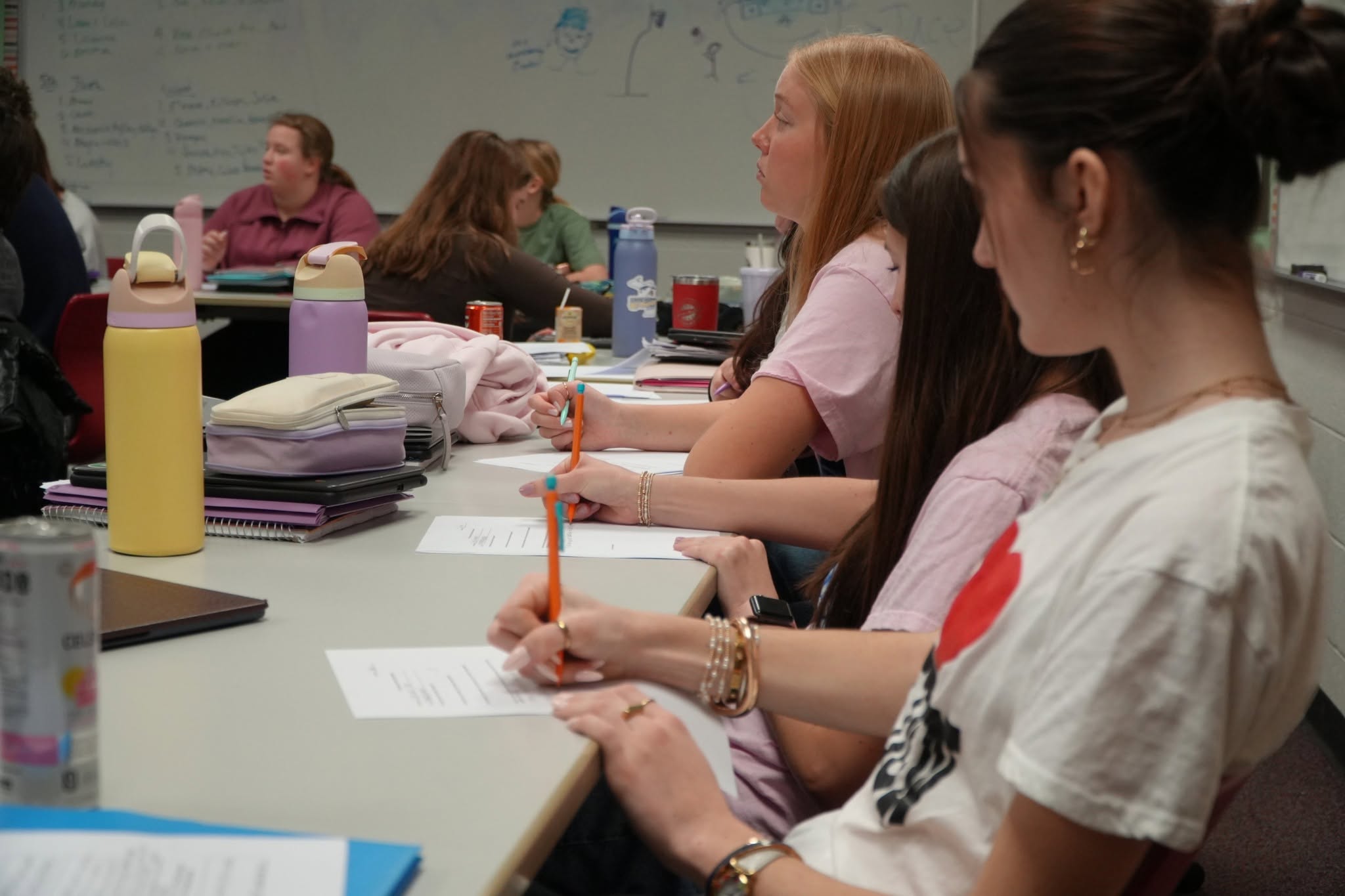 Students working at a long classroom table with worksheets and pencils